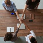 A diverse group of professionals engaging in a teamwork celebration with a fist bump over a wooden table indoors.